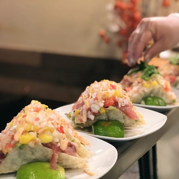 Three Asian-fusion tacos with diced toppings and lime wedges on white plates, with a hand garnishing one.