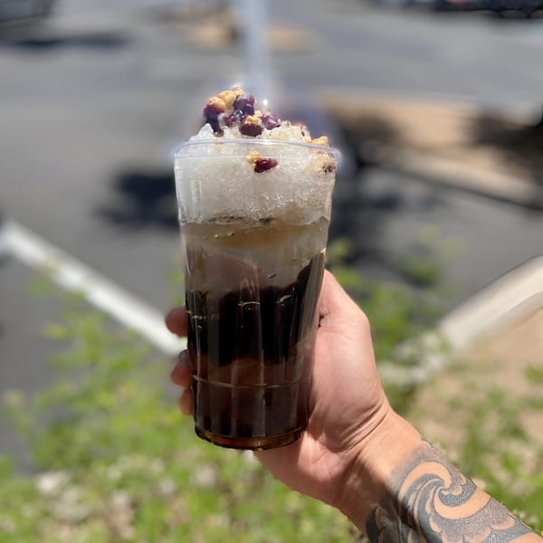A hand with a tattooed arm holds a clear cup of iced tea topped with fruit and nuts.