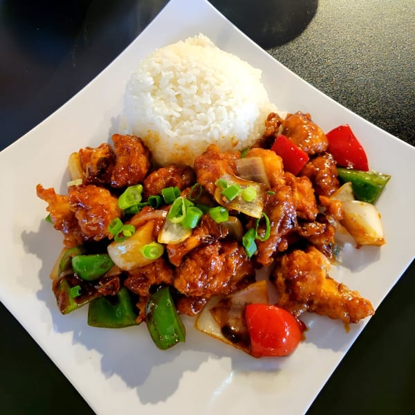 A white plate of orange chicken with white rice, green onions, and bell peppers.